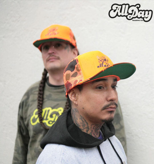 Two people wearing colorful sweatshirts and caps with 'All Day' branding against a plain background.