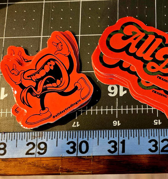 Two red and black All Day stickers on a ruler for size reference